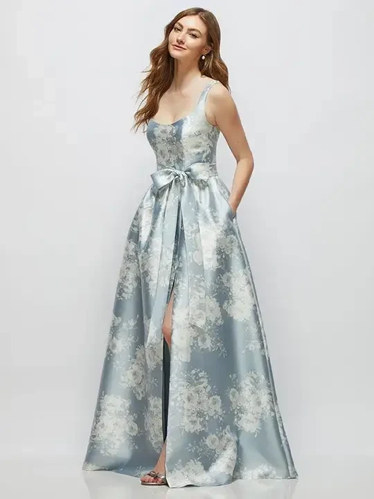 A woman in a sleeveless, floral-patterned blue gown with a bow at the waist, posing with her hands in the pockets.