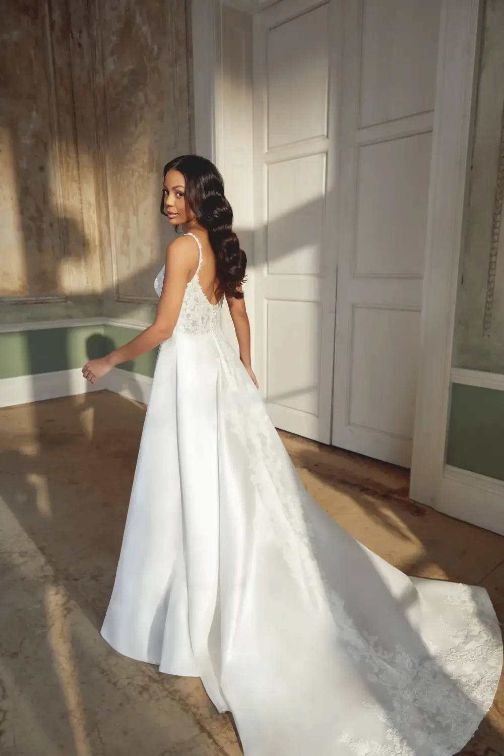 A woman wearing a white wedding dress, standing with her back to the camera, showcasing the dress's train and lace detailing. The setting features elegant interior decor with natural light.