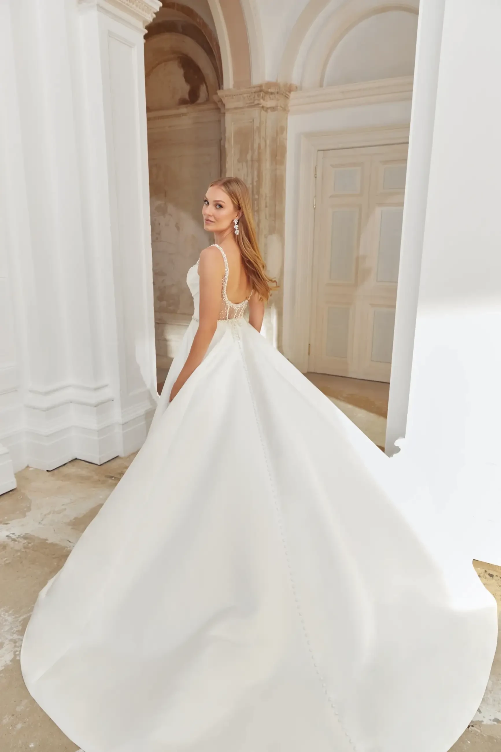 A woman in a luxurious white gown stands gracefully, showcasing the back of the dress. The setting features elegant architecture with soft, warm lighting.