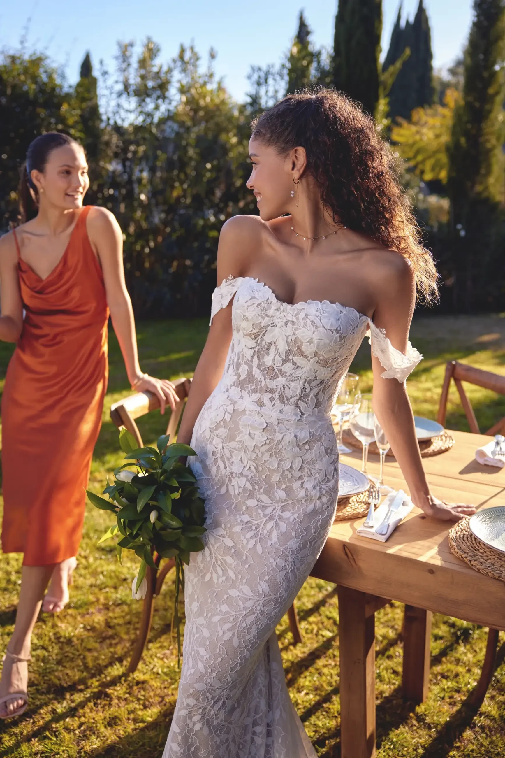 A bride in a white lace dress with a bouquet stands by a sunlit outdoor table. A smiling woman in an orange dress is beside her, creating a joyful scene.