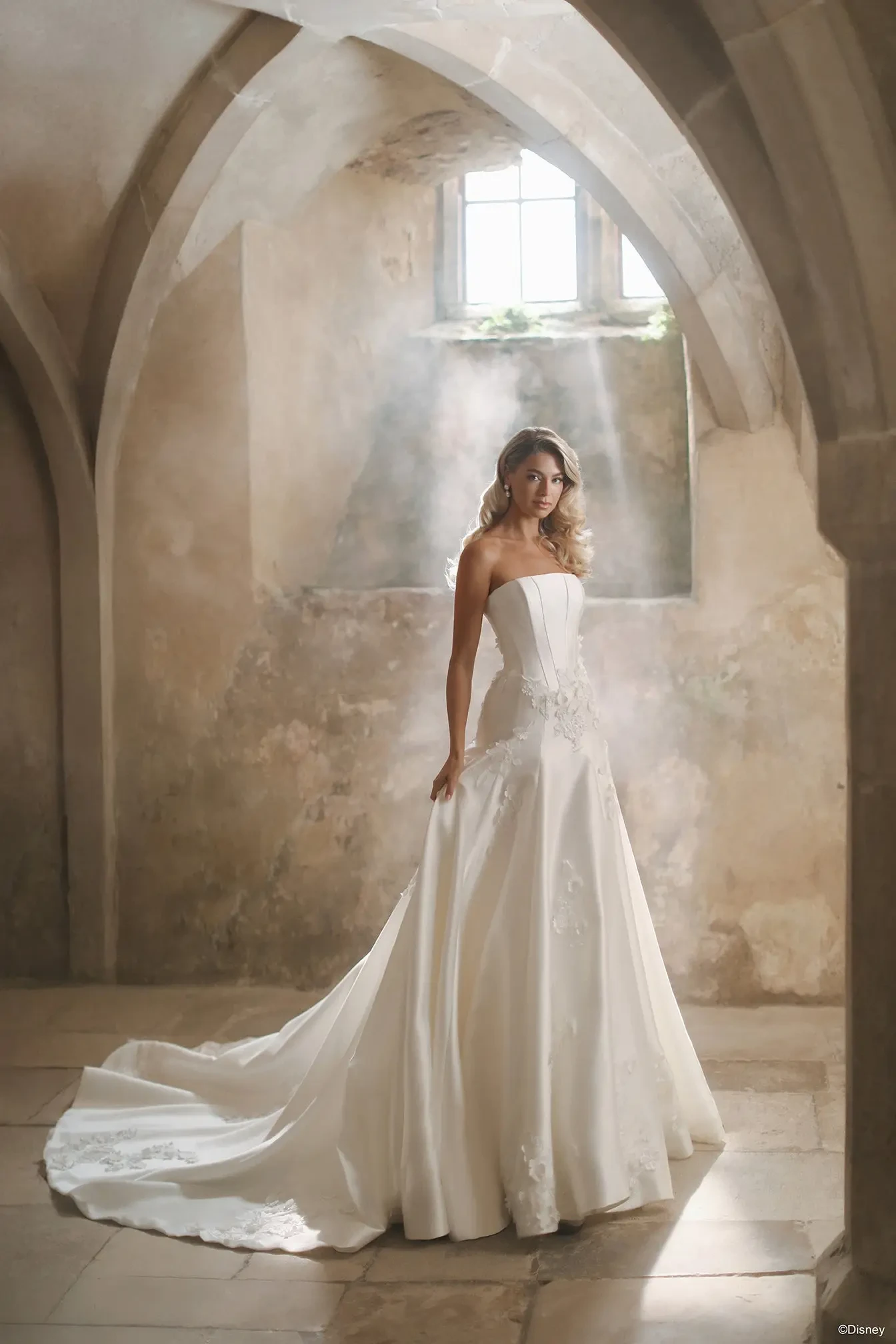 A woman in a strapless white wedding gown stands gracefully in a softly lit, stone-walled room, with light filtering through a window and creating an ethereal atmosphere.