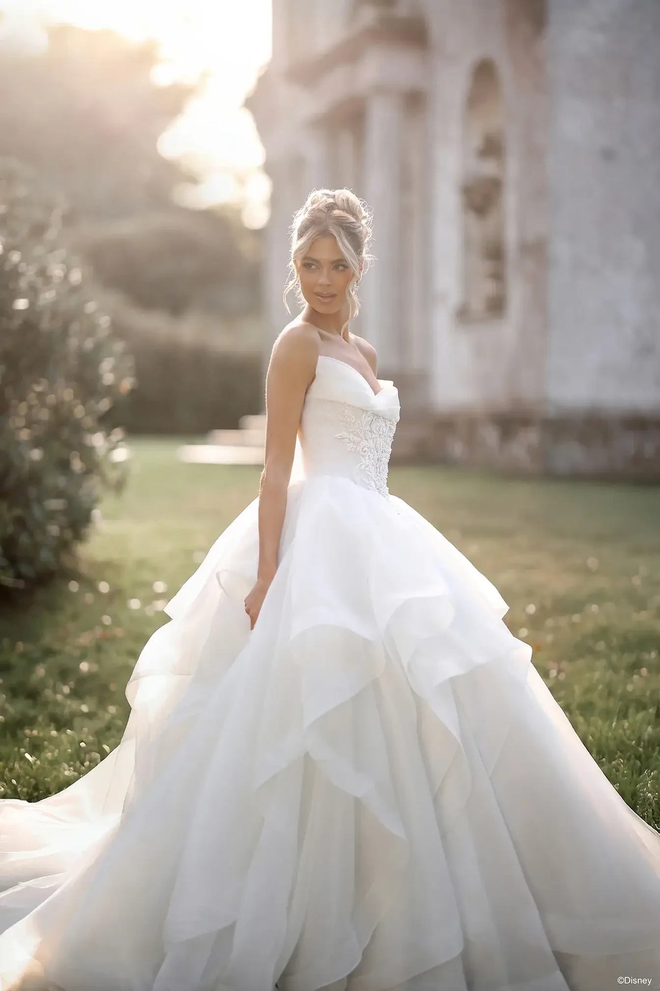 A woman in a radiant white wedding dress stands gracefully outdoors, with soft sunlight illuminating her figure. She has an elegantly styled hairdo and is looking over her shoulder at the camera.