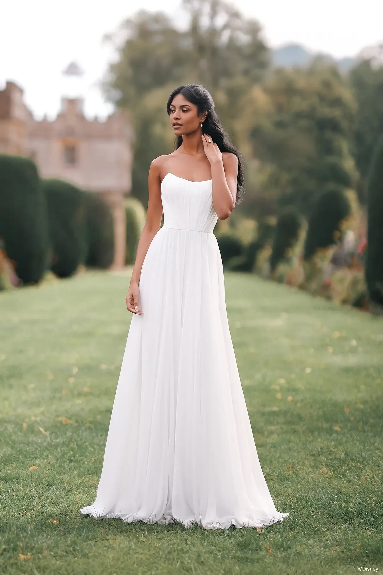 A woman in a white, floor-length gown stands elegantly on green grass, with manicured hedges and a blurred background of trees and architecture.