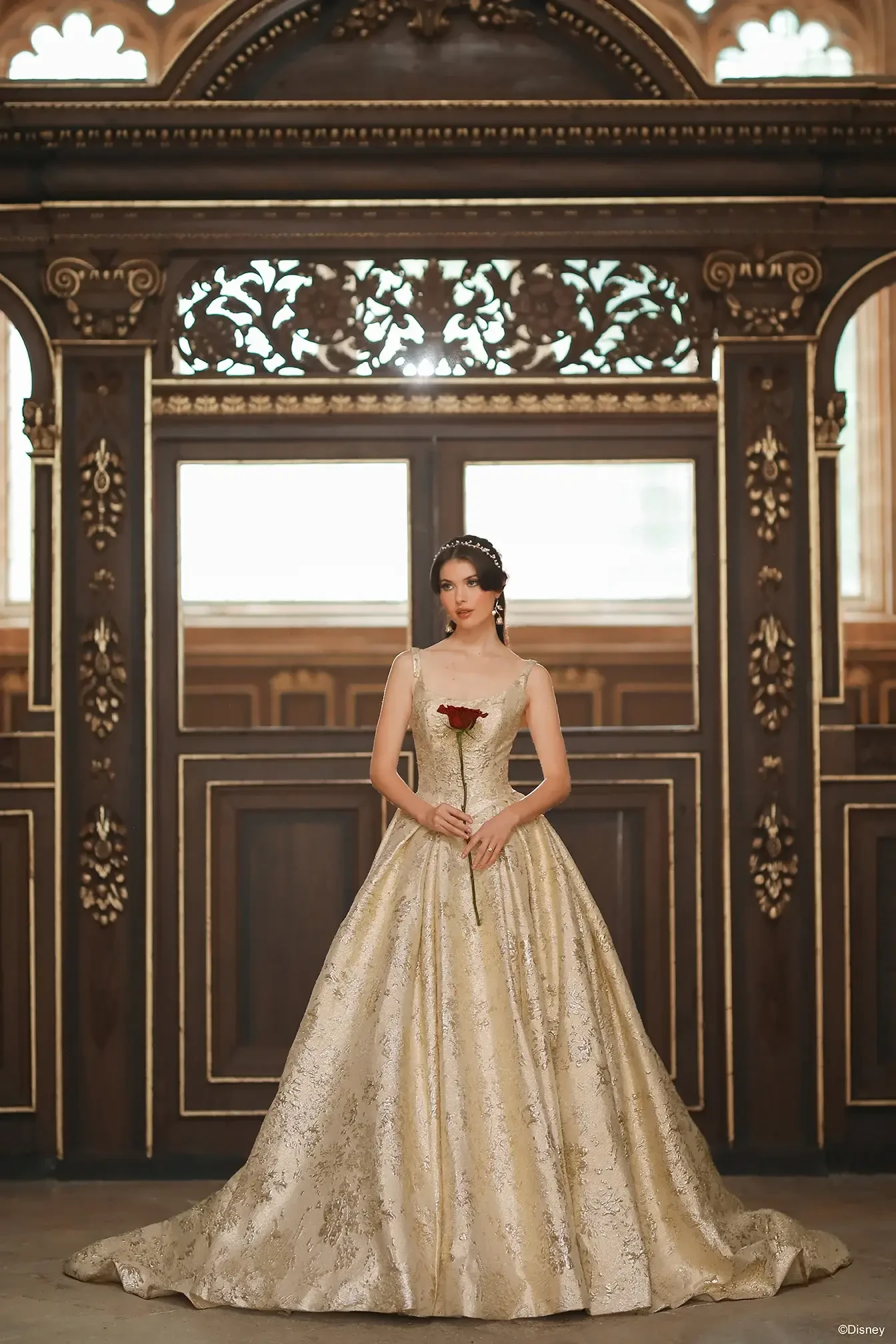 A woman in an elegant, gold gown holding a rose, standing in a grand, ornately decorated room with wooden details.