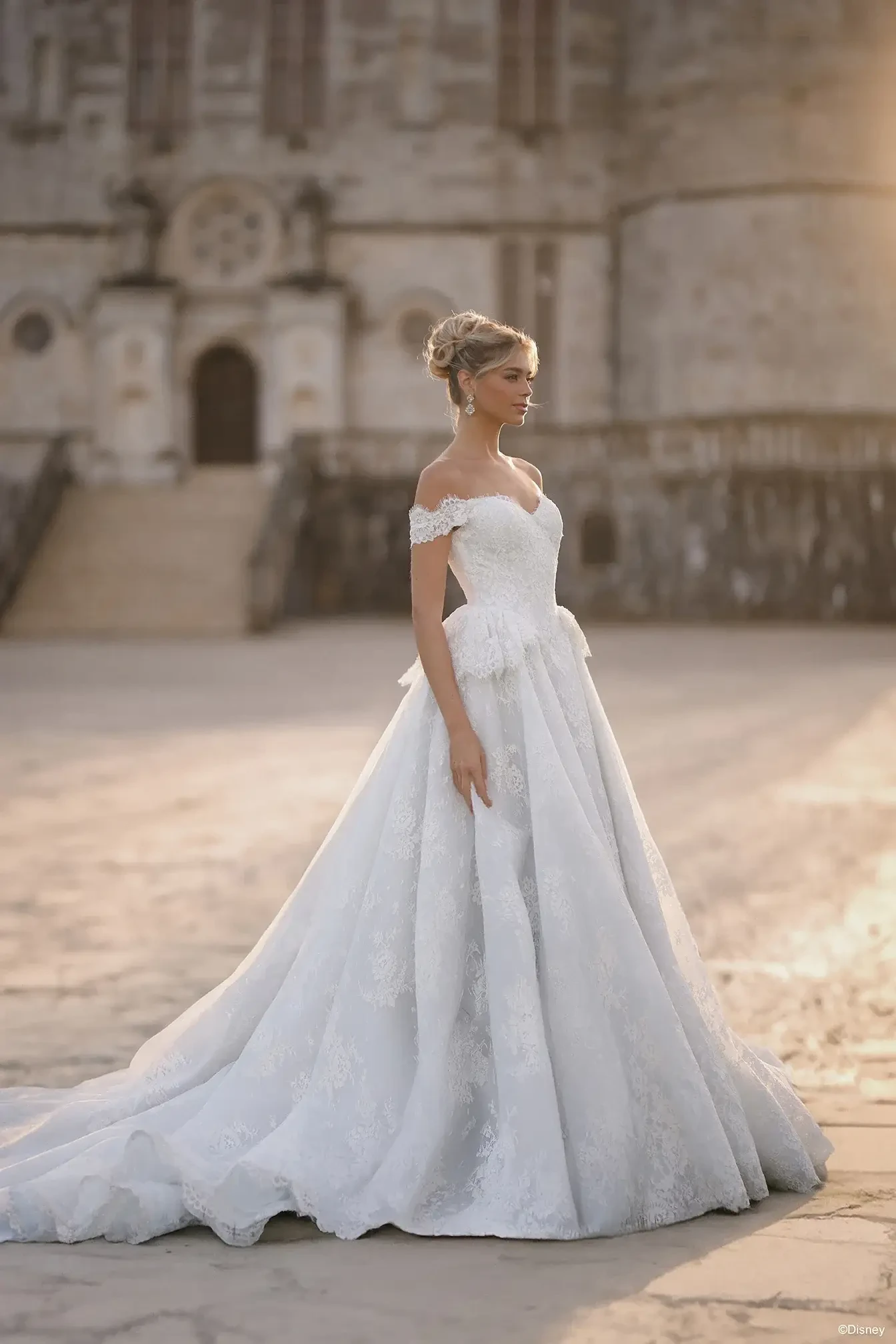 A woman in a beautiful off-the-shoulder white wedding dress stands on a stone pathway with an ornate building in the background, illuminated by soft sunlight.