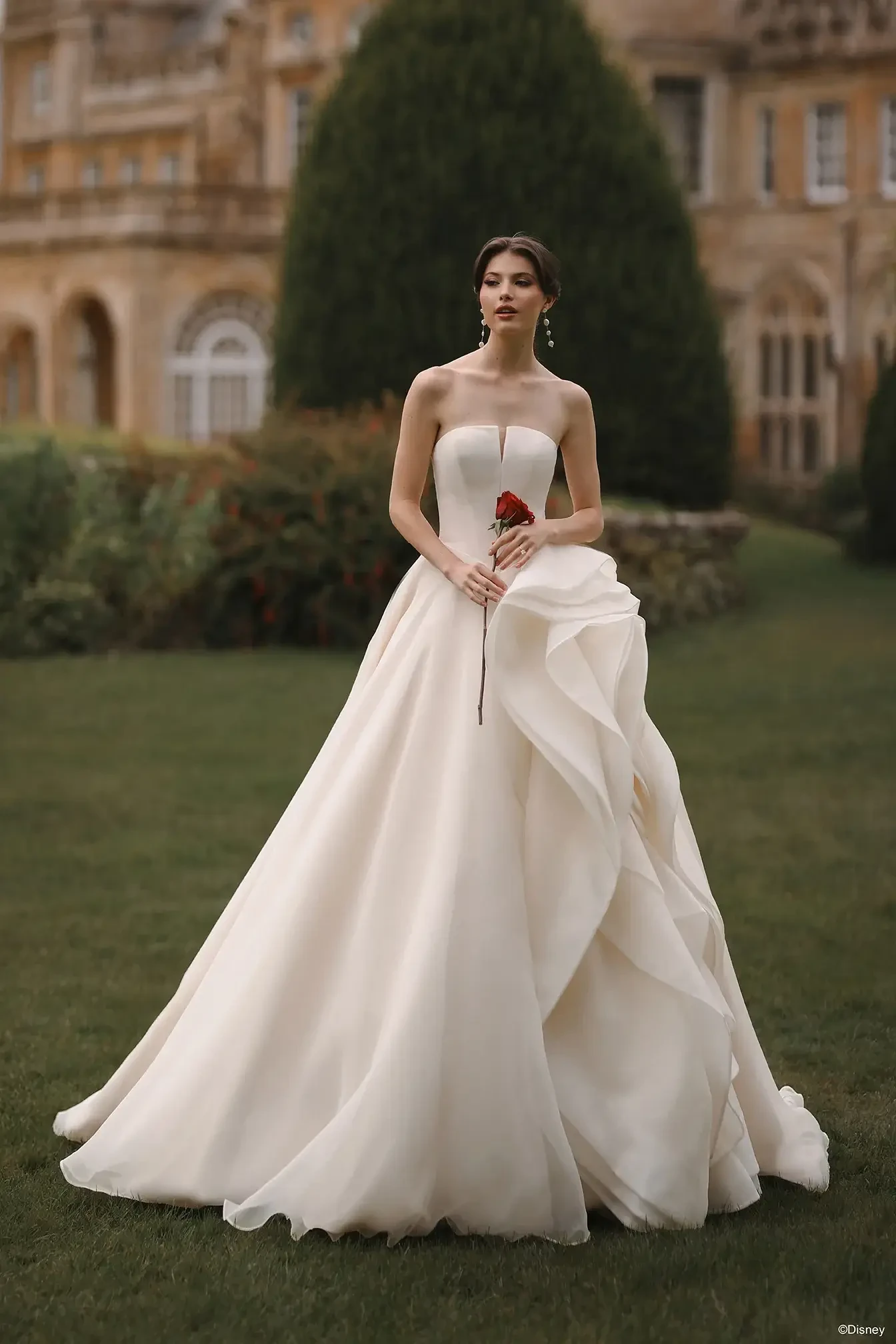 A woman in a strapless white wedding gown holding a red rose, standing in a garden with an elegant building in the background.