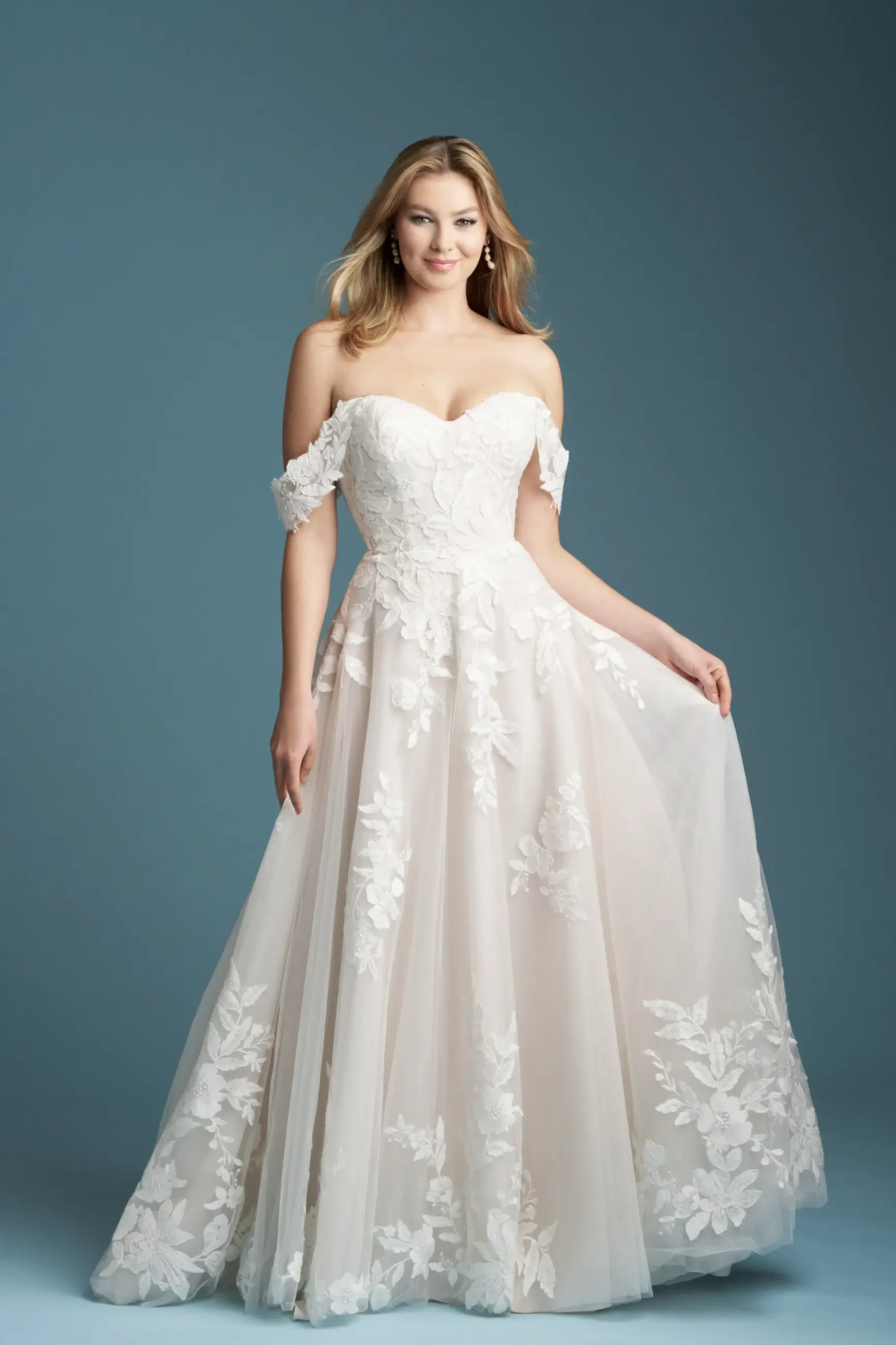 Model is wearing an A-line gown with a sweetheart neckline, airy off-the-shoulder sleeves, and sparkle tulle adorned with bold lace appliques.