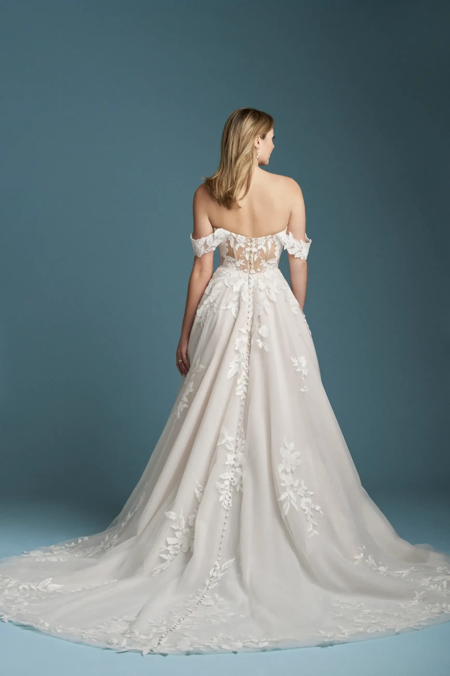 Model is wearing an A-line gown with a sweetheart neckline, airy off-the-shoulder sleeves, and sparkle tulle adorned with bold lace appliques.