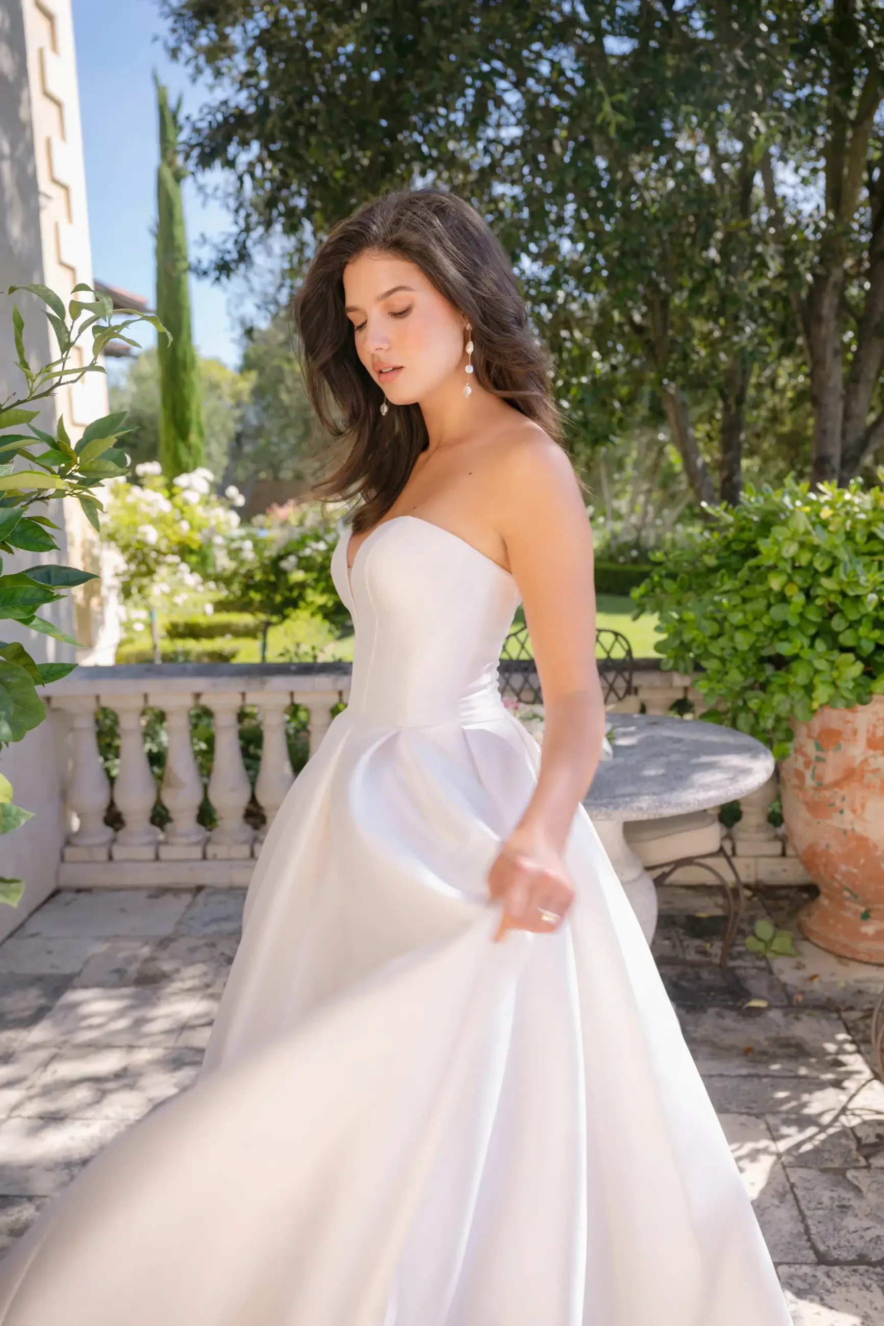 Brunette woman in a strapless wedding gown stands on a sunny patio, surrounded by lush greenery. She looks serene and elegant, holding her dress.