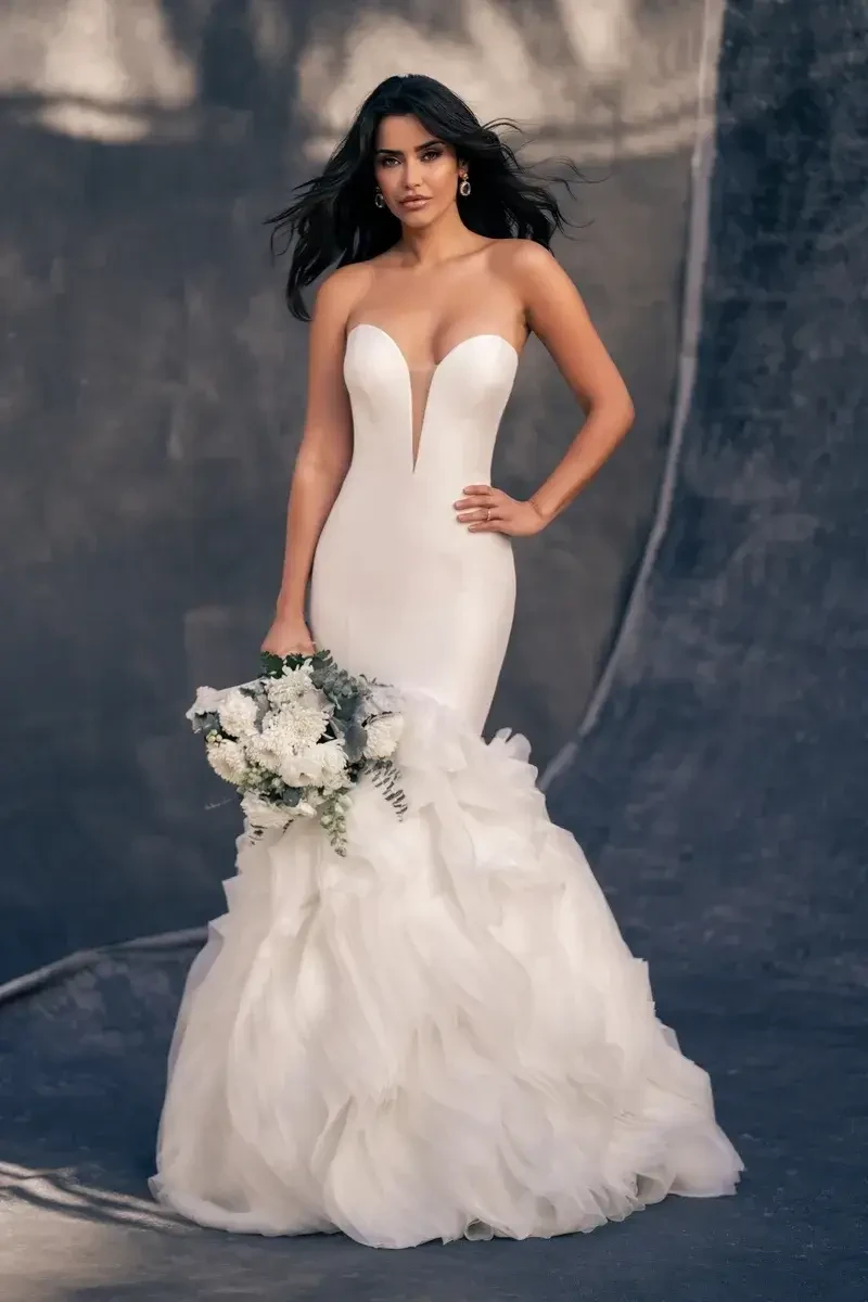 A woman wearing a stunning white wedding dress with a fitted bodice and a ruffled skirt, holding a bouquet of flowers.