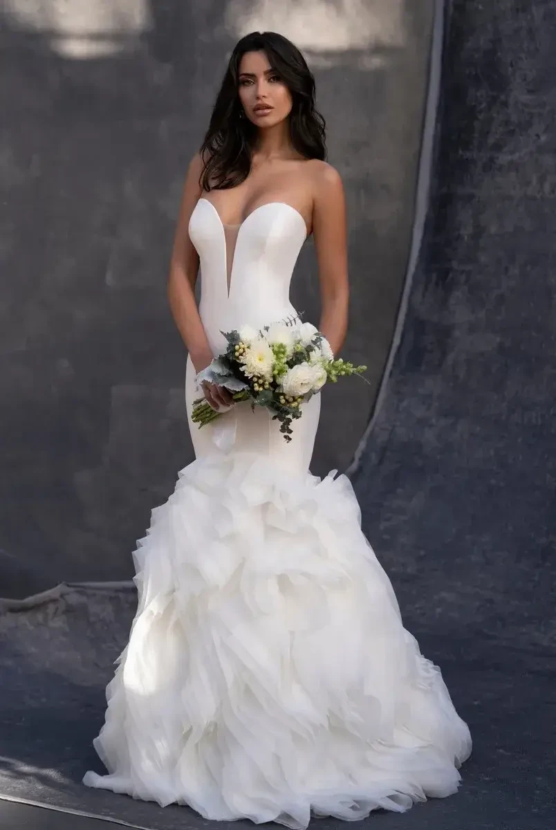 A woman wearing a stunning white wedding dress with a fitted bodice and a ruffled skirt, holding a bouquet of flowers.