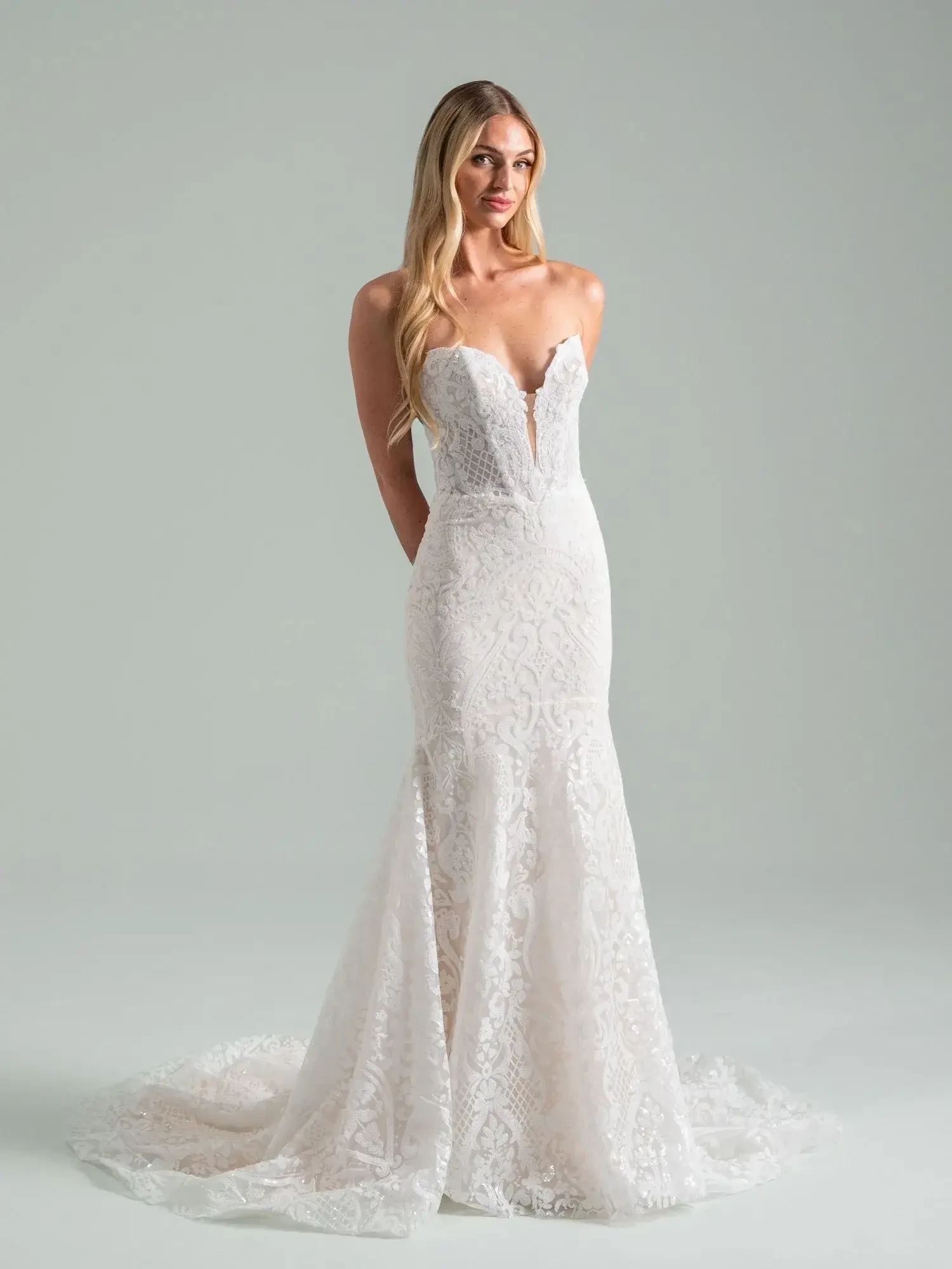 A model wearing a strapless, white wedding gown with intricate lace details standing against a light gray background.