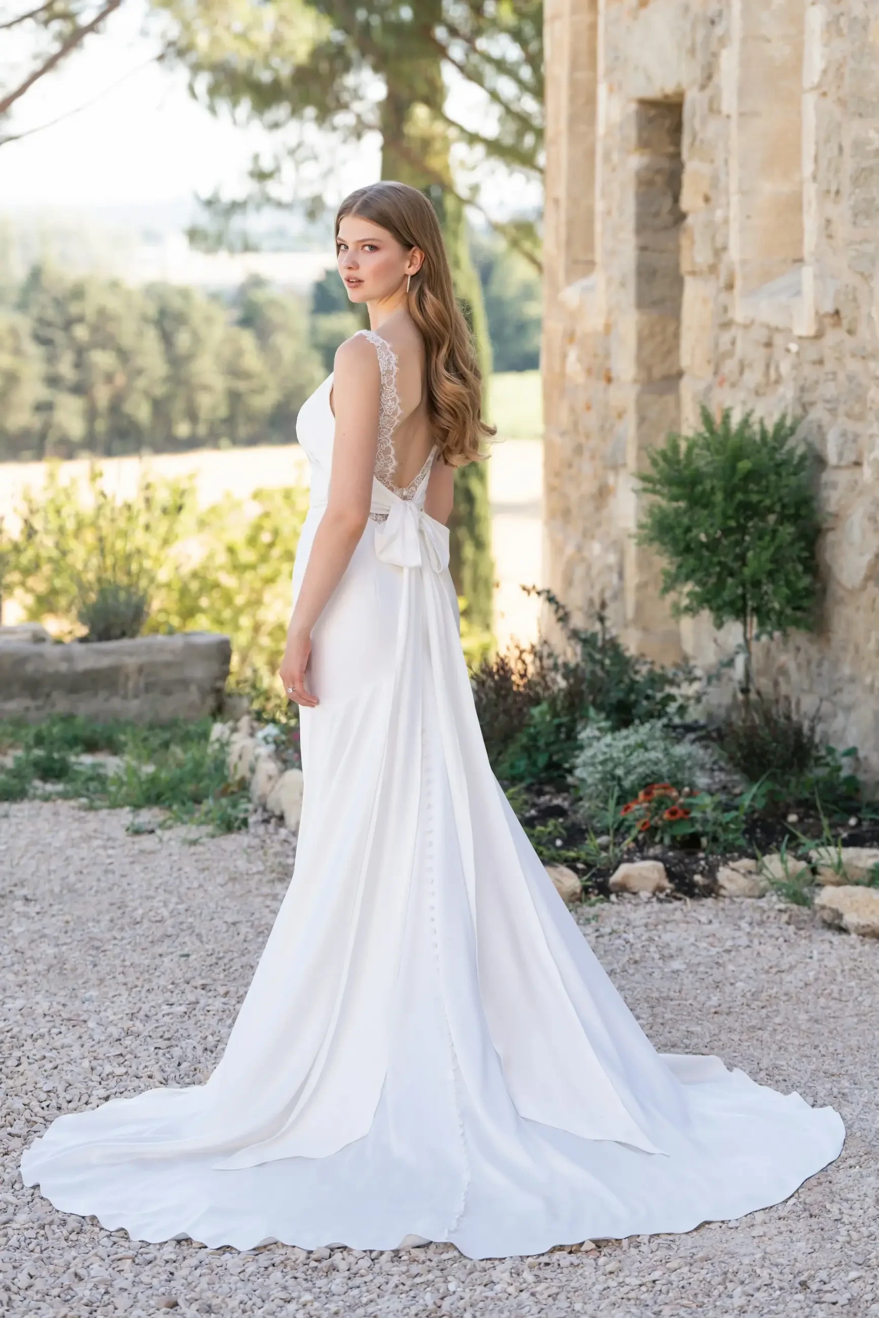 Model is wearing a stretch satin sheath wedding dress with lace accents and a tie back
