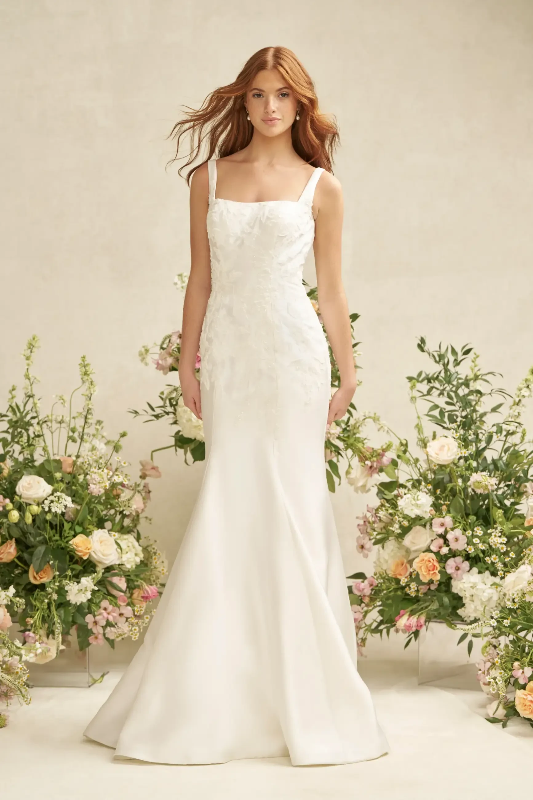 A woman in a fitted, elegant white gown stands confidently amid lush floral arrangements. The scene conveys a serene, bridal elegance.