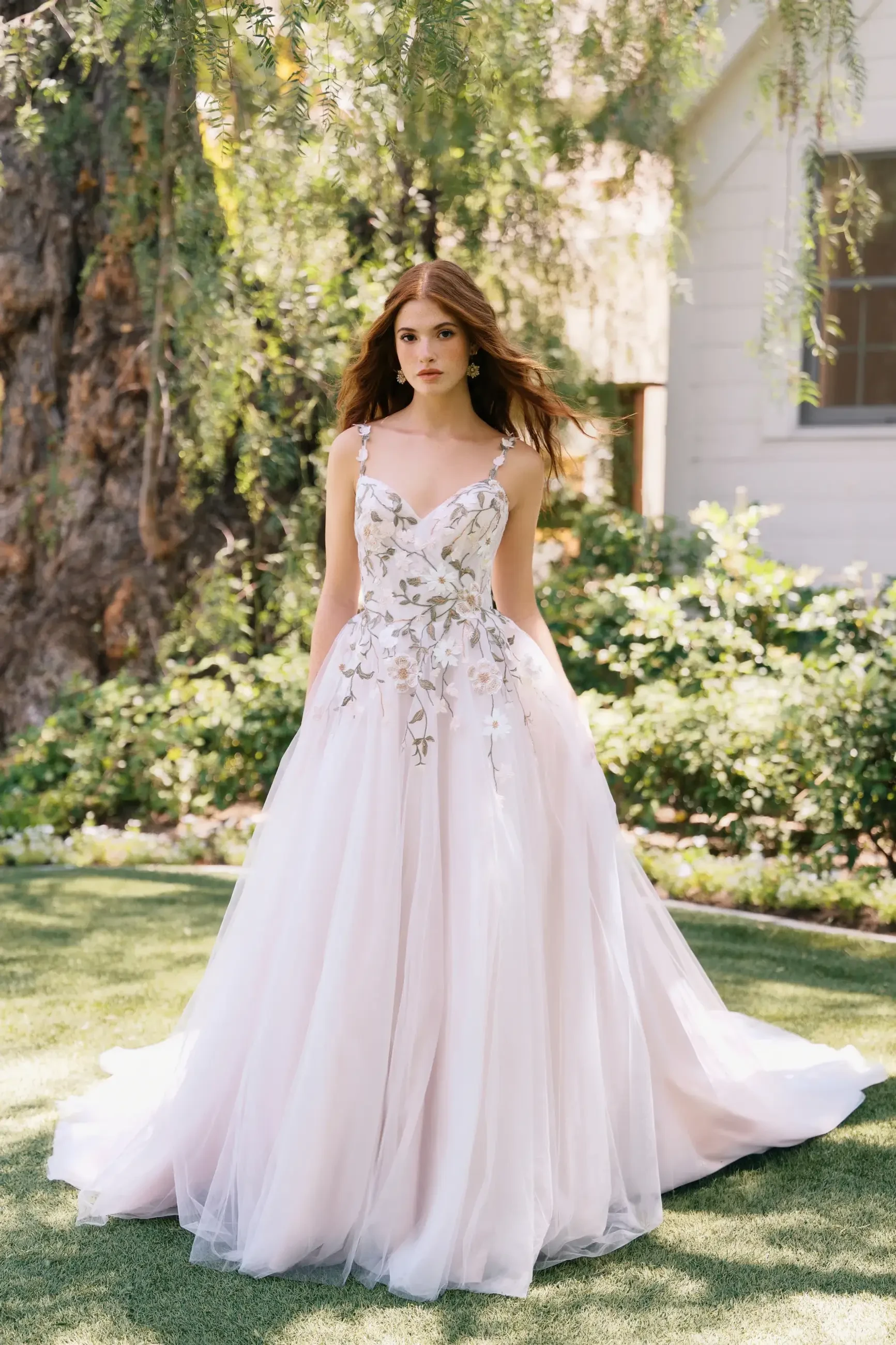 A woman in a flowing, embellished gown stands in a garden with greenery and trees in the background.
