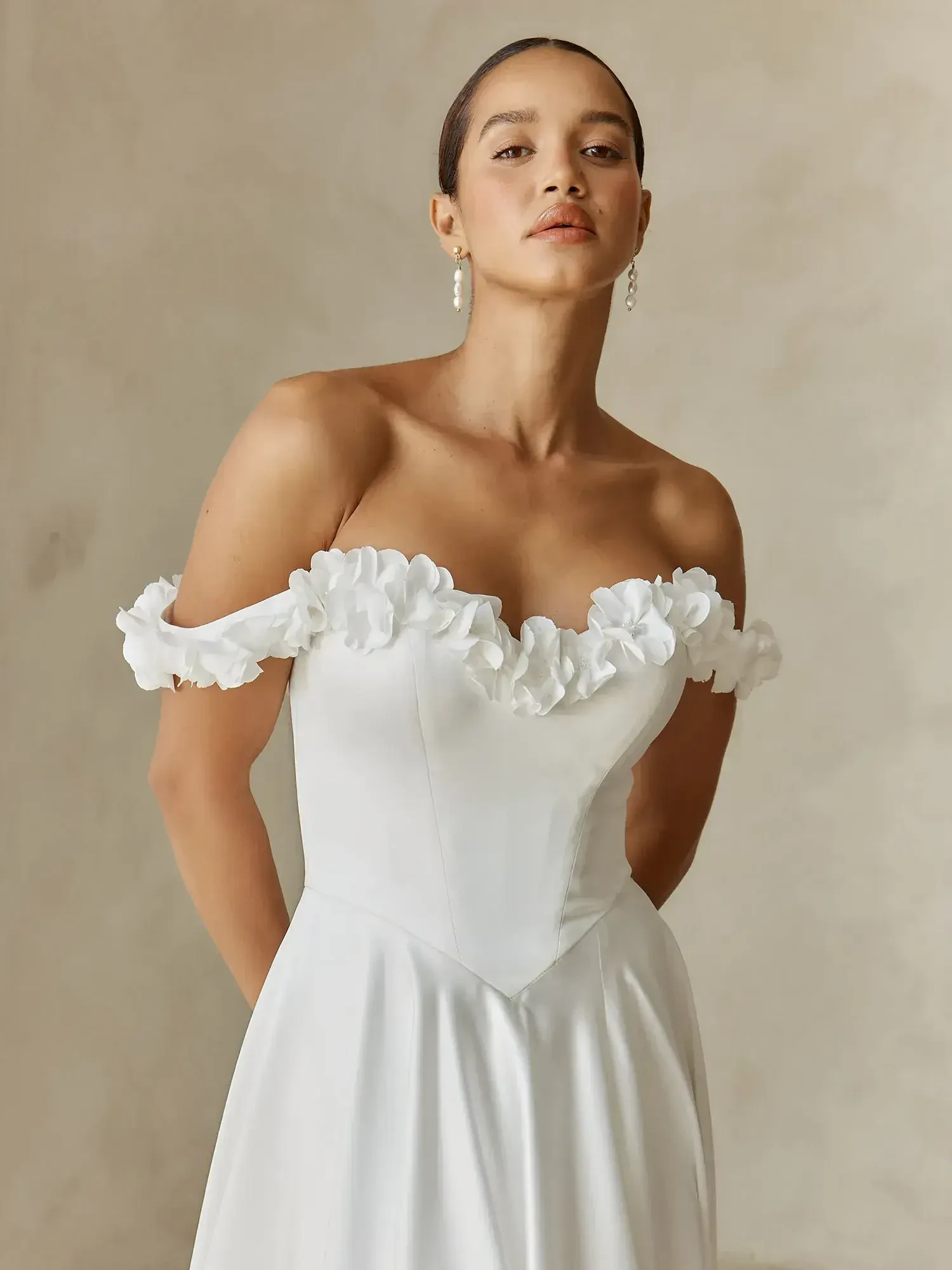 A woman in a white dress with off-the-shoulder sleeves adorned with floral embellishments, standing against a neutral background.