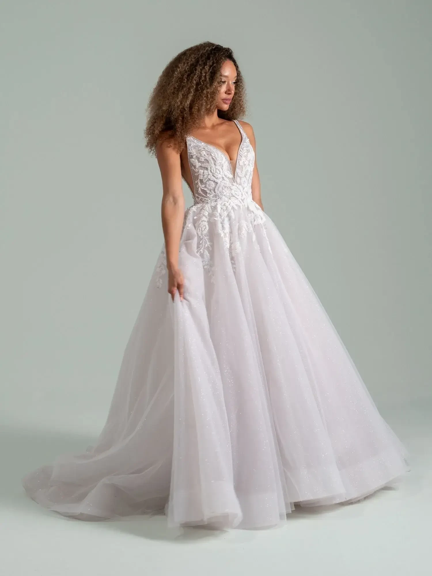 A woman wearing a white bridal gown with intricate lace details and a flowing skirt. She has curly hair and is standing elegantly, holding part of the skirt.